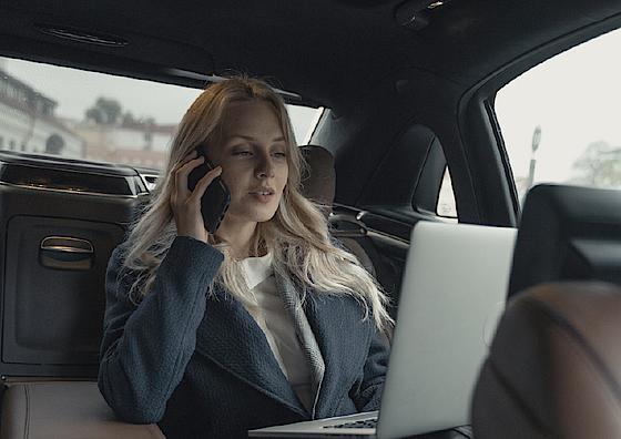 Eine junge Frau sitzt im Fond eines Autos und telefoniert. Auf ihrem Schoß steht ein aufgeklappter Laptop.